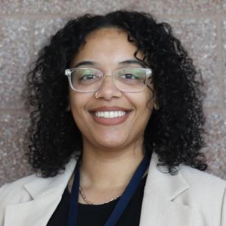 Headshot of Bryanna, a female-presenting person with medium skin tone, dark curly hair, wearing glasses and a white suit and black t-shirt