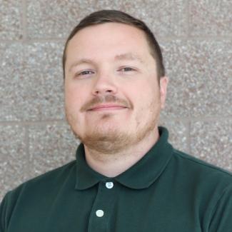 Headshot of Tanner, a male-presenting person with light skin, close-cropped brown hair and stubbly beard, wearing a dark green polo shirt.